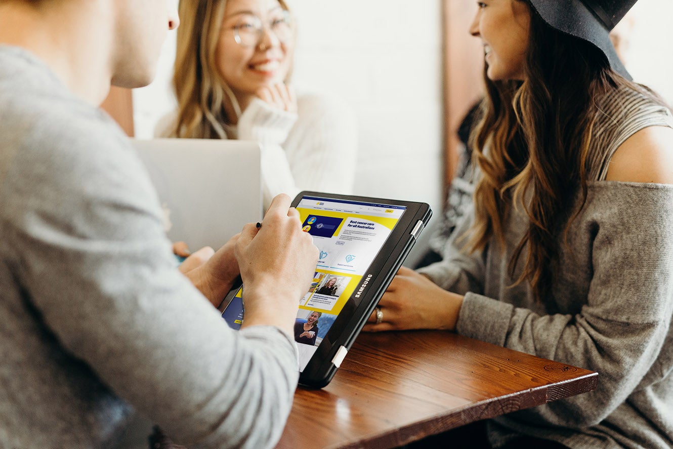 Three people smiling, one is holding an iPad showing a Cancer Council page and another has a laptop open.